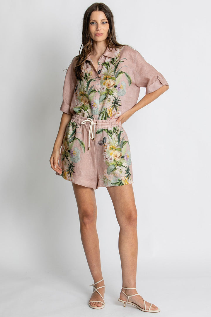 A woman modeling a dusty pink short-sleeve romper with a green and yellow tropical floral print, featuring a drawstring waist and matching shorts, paired with beige strappy sandals against a light gray studio background.
