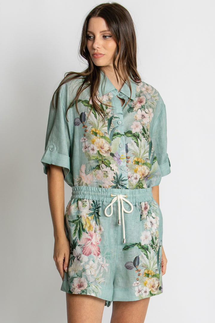 A woman modeling a mint green short-sleeve button-up shirt and matching shorts set with a colorful floral print, featuring a white drawstring at the waist, against a light gray studio background.