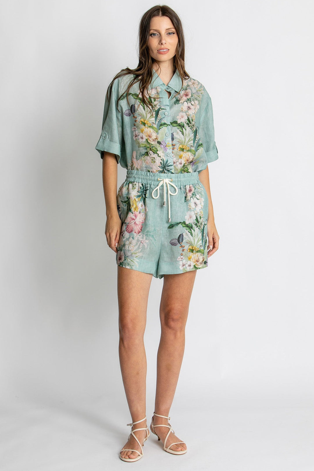A woman modeling a matching two-piece set consisting of a short-sleeve button-up shirt and coordinating shorts, both in a light sage green color with a colorful floral and butterfly print. She is wearing beige strappy sandals against a plain light gray studio background.