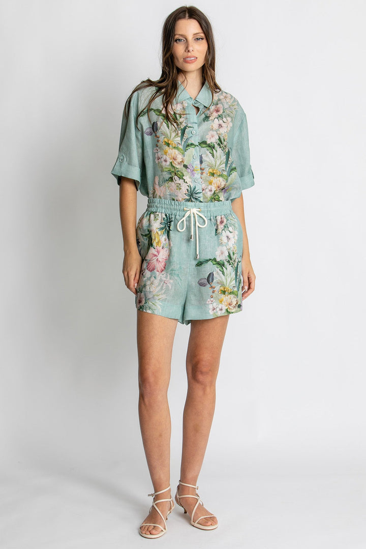 A woman modeling a matching two-piece set consisting of a short-sleeve button-up shirt and coordinating shorts, both in a light sage green color with a colorful floral and butterfly print. She is wearing beige strappy sandals against a plain light gray studio background.