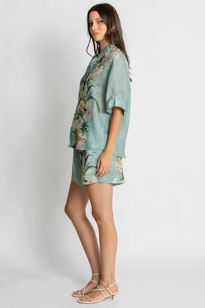 A woman modeling a matching light blue-green short-sleeve button-up shirt and shorts set with a floral print, paired with beige strappy sandals, against a plain light gray studio background.