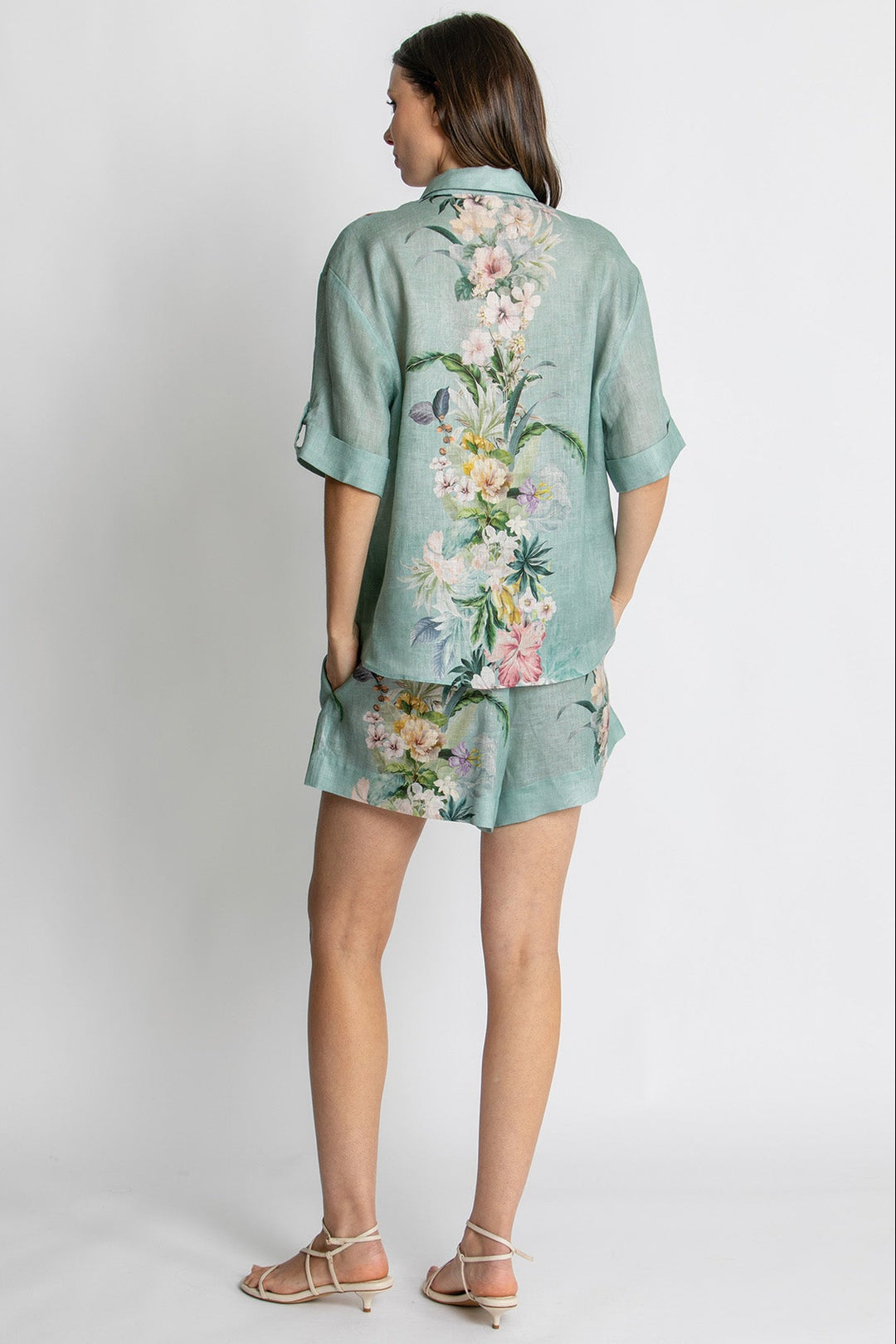 A woman shown from the back wearing a mint green short-sleeve button-up shirt and matching shorts set with a large multicolor floral print, paired with beige strappy sandals, against a light gray studio background.
