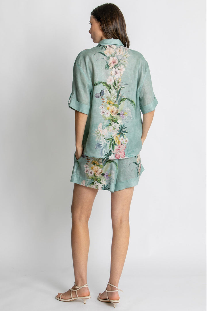 A woman shown from the back wearing a mint green short-sleeve button-up shirt and matching shorts set with a large multicolor floral print, paired with beige strappy sandals, against a light gray studio background.