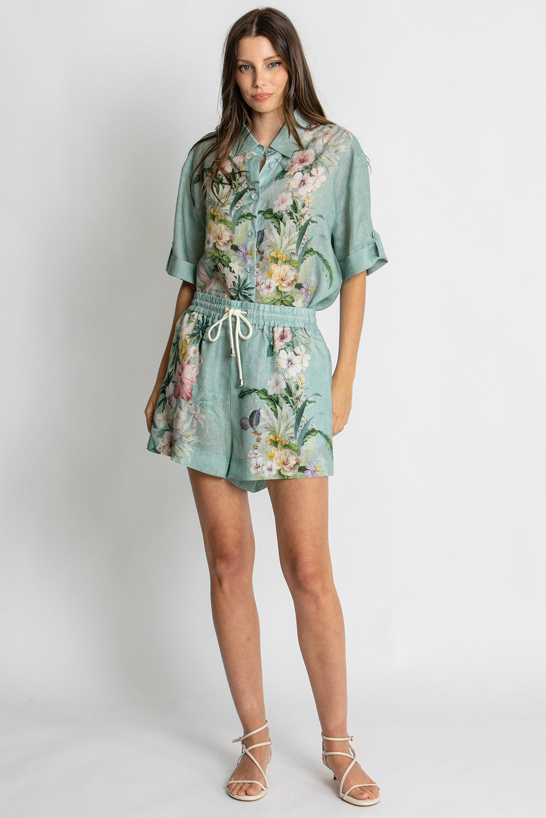 A woman modeling a sage green short-sleeve romper with a colorful floral print, featuring a white drawstring waist, against a light gray studio background.
