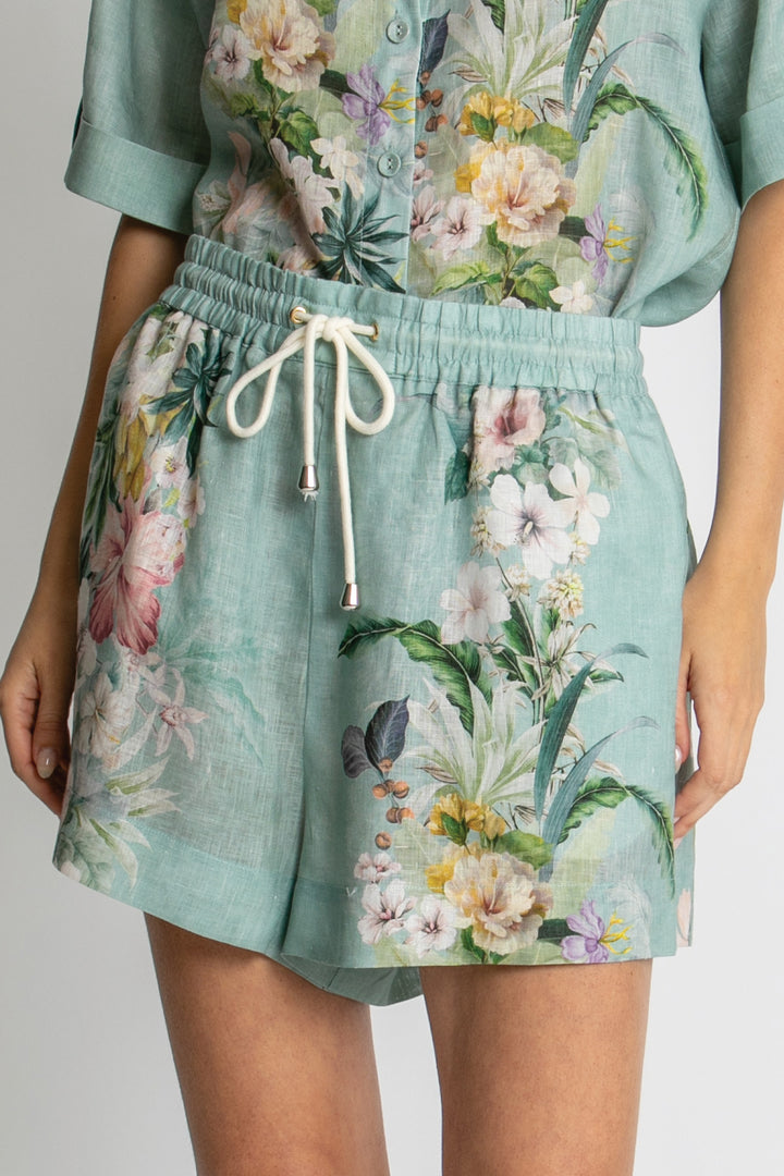 A close-up of a person wearing mint green floral shorts with a white drawstring and a matching floral top, shown against a light gray studio background.