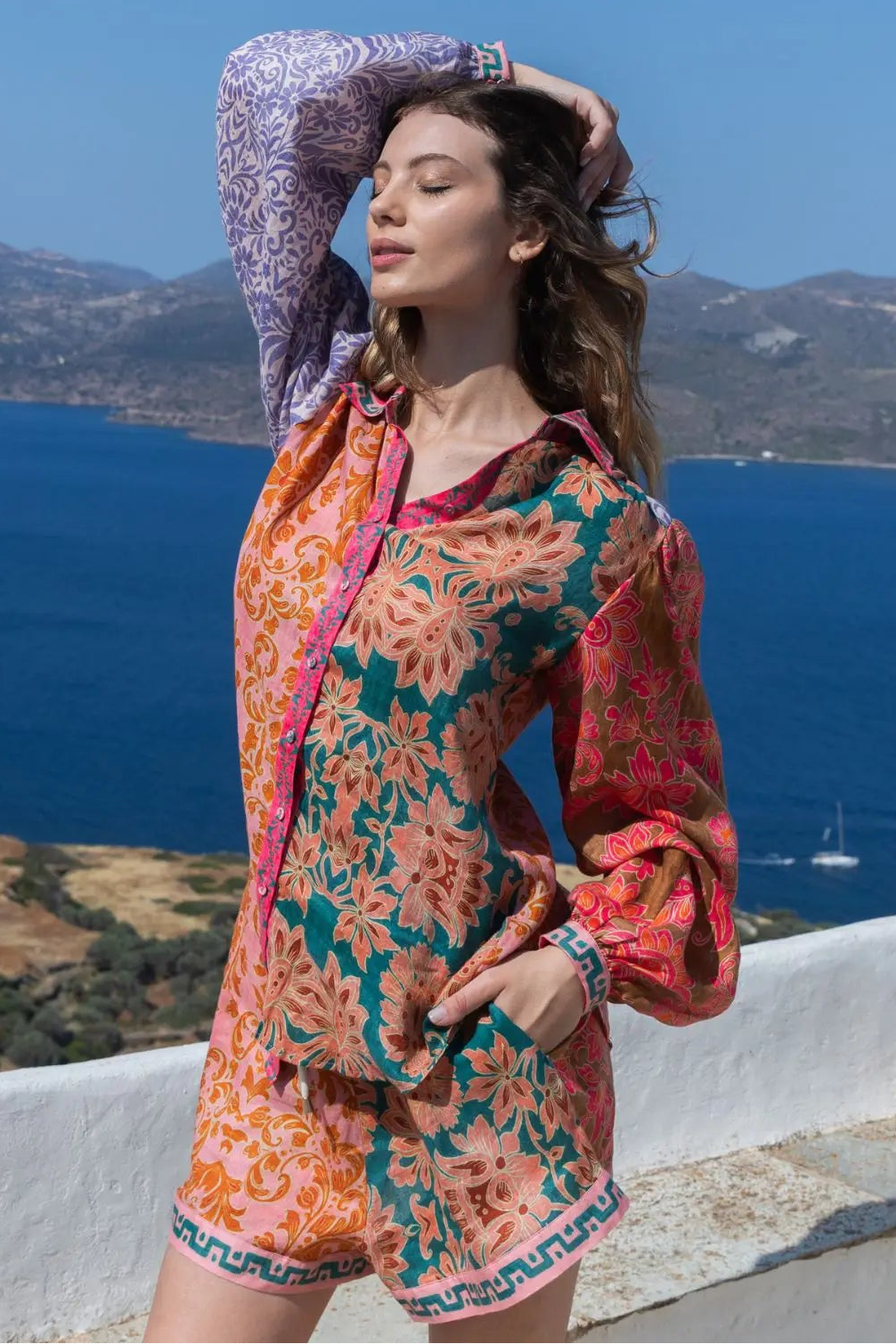 Woman in a colorful floral shirt & shorts standing against a scenic backdrop of blue sea and mountains.
