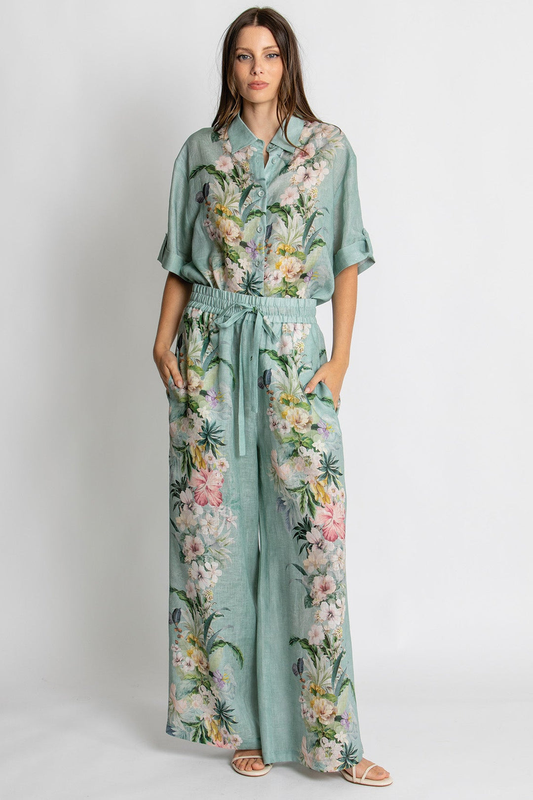 A woman modeling a mint green two-piece set featuring a short-sleeve button-up shirt and matching wide-leg pants, both decorated with a colorful tropical floral and palm leaf print. She is standing against a plain light gray studio background.