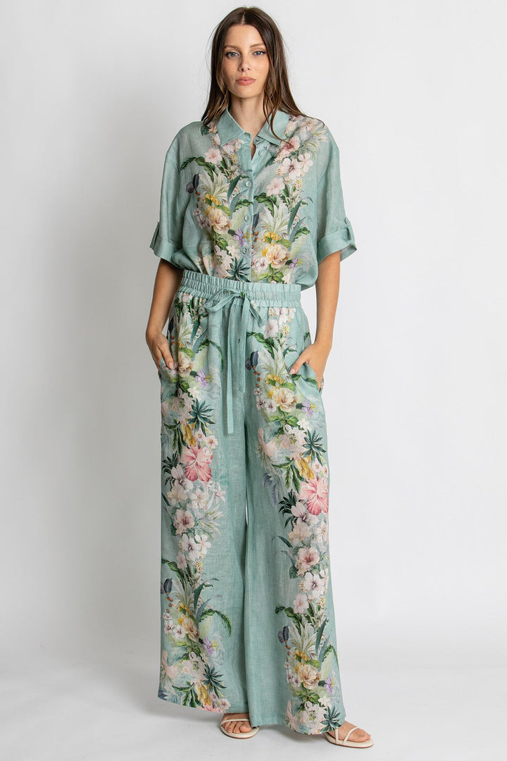 A woman modeling a mint green two-piece set featuring a short-sleeve button-up shirt and matching wide-leg pants, both decorated with a colorful tropical floral and palm leaf print. She is standing against a plain light gray studio background.