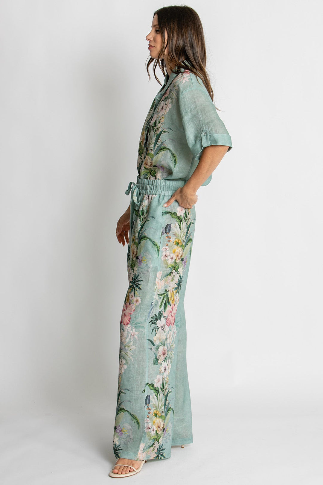 A woman modeling a sage green floral print jumpsuit with short sleeves, a tie waist, and wide-leg pants, shown from a side profile against a light gray studio background.