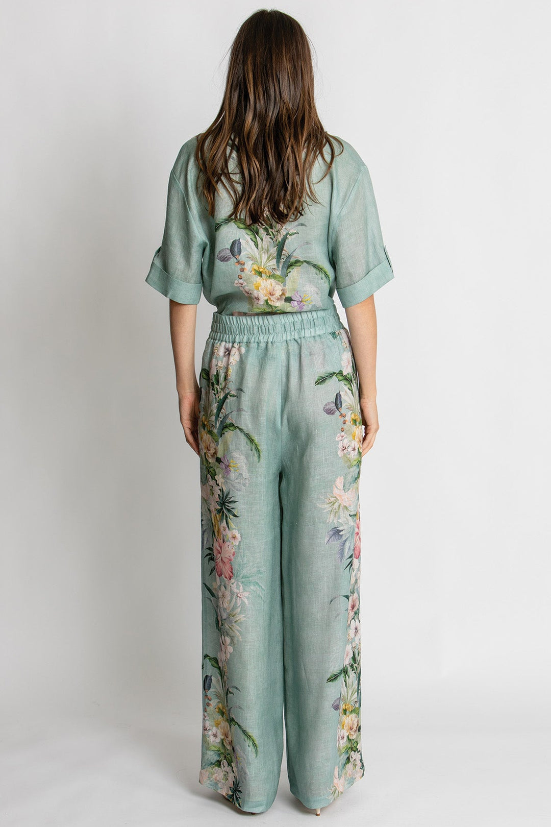 A woman viewed from behind wearing a sage green jumpsuit with a floral print, standing against a plain light gray studio background.