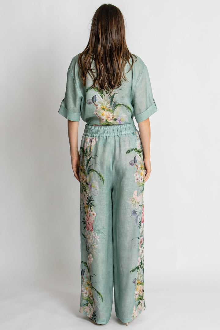 A woman viewed from behind wearing a sage green jumpsuit with a floral print, standing against a plain light gray studio background.