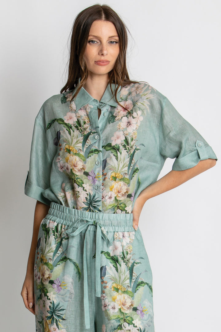 A woman modeling a matching two-piece set consisting of a short-sleeve button-up shirt and coordinating drawstring pants, both in a light sage green fabric with a colorful tropical floral and bird print.