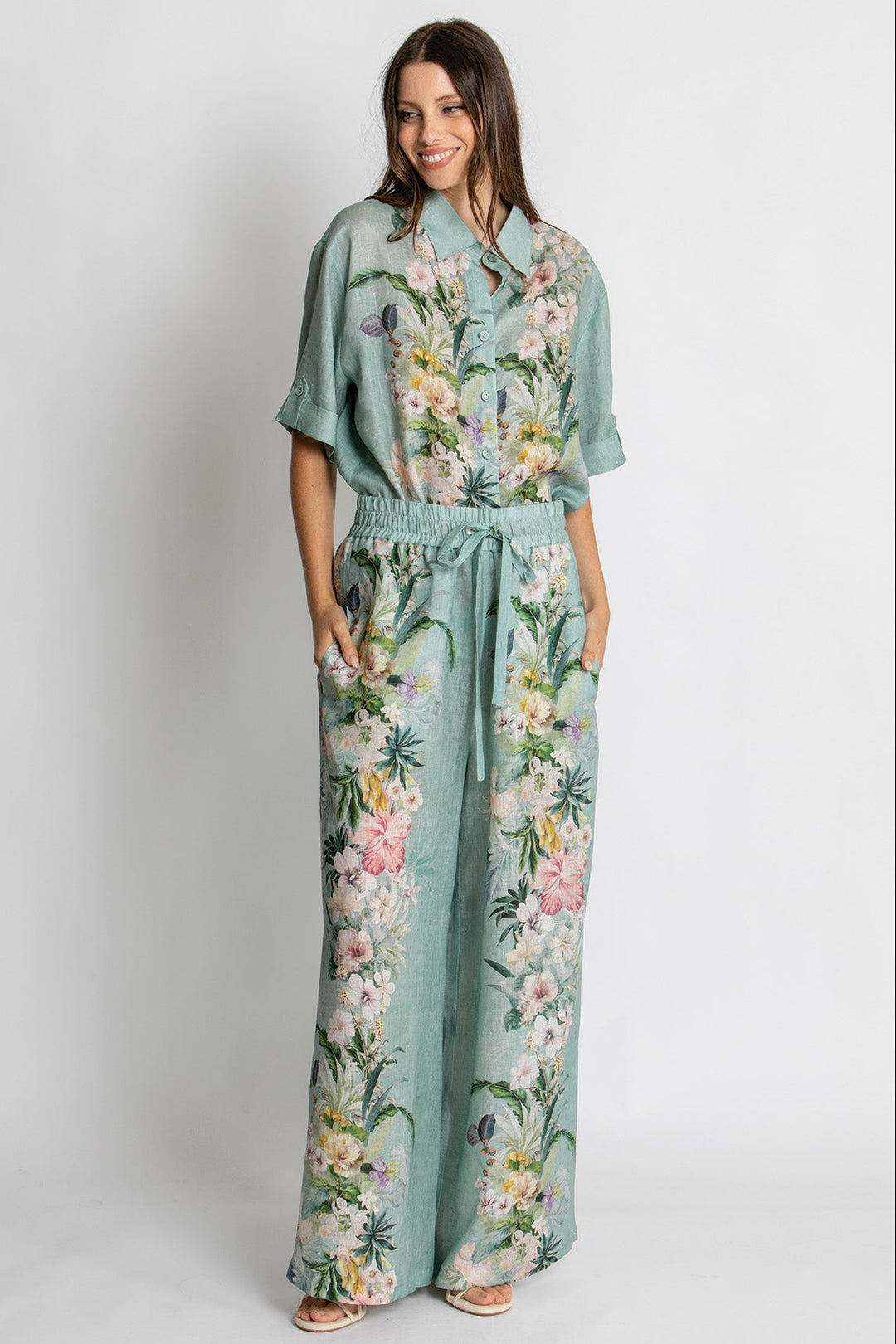 A woman modeling a matching two-piece set consisting of a short-sleeve collared top and wide-leg pants, both in a sage green color with a vibrant multicolor floral print, set against a plain light gray background.