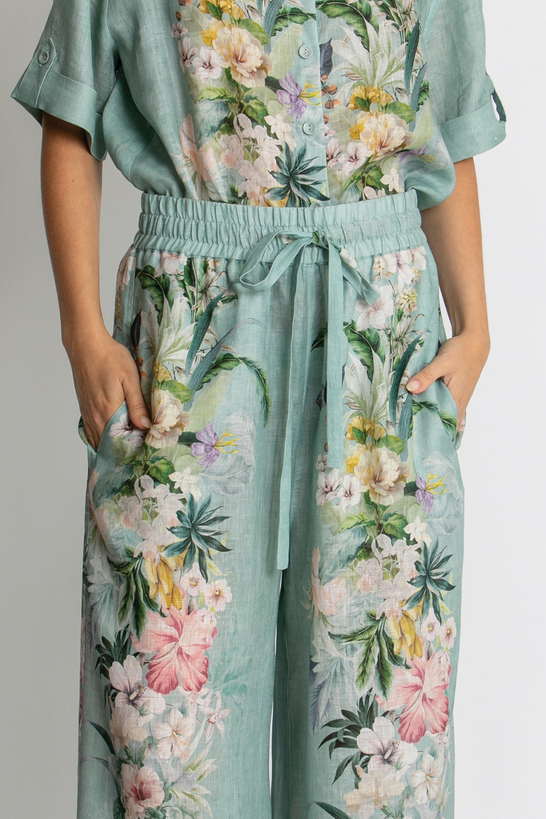 A person wearing a mint green floral jumpsuit with a tie waist and side pockets, shown from the chest down against a plain light background.
