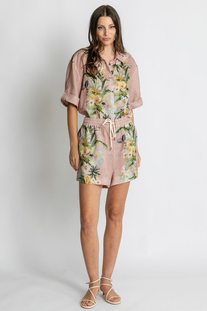 A woman modeling a short-sleeve romper with a dusty pink base and a tropical floral print, paired with beige strappy sandals against a light gray studio background.
