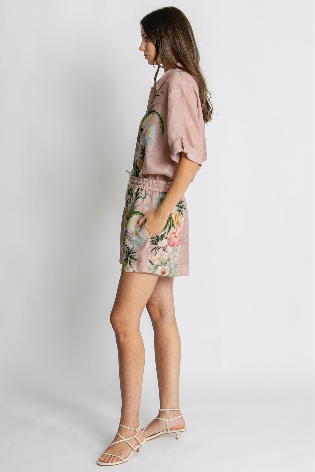 A woman modeling a dusty pink short-sleeve top and matching floral-print shorts against a light gray studio background.