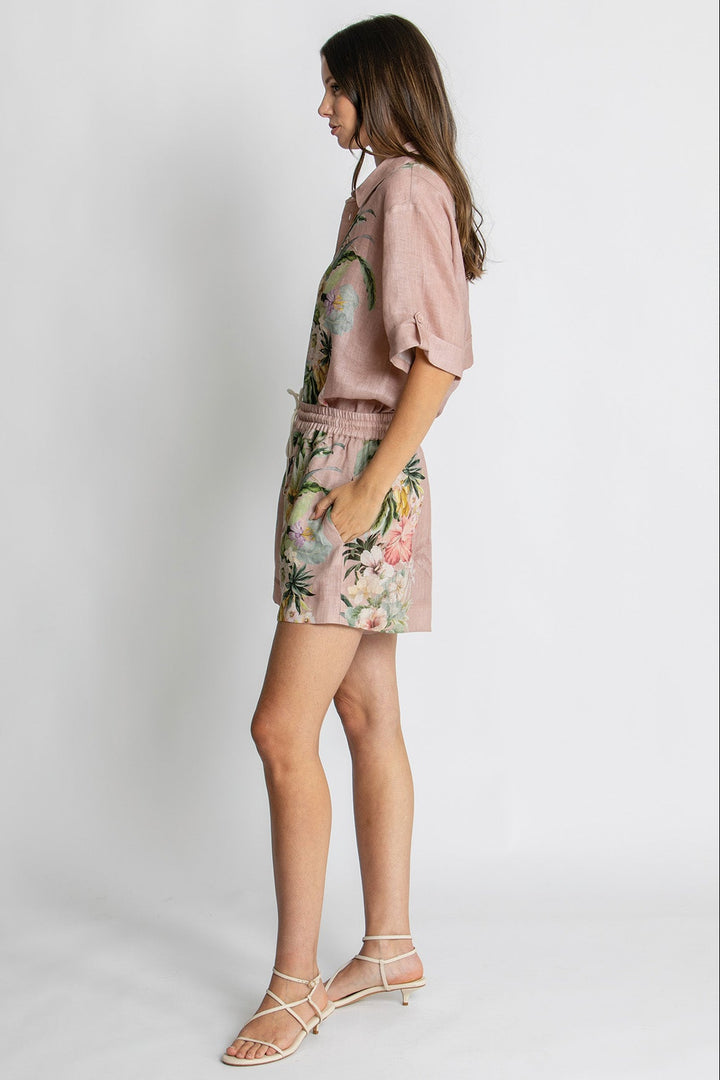 A woman modeling a dusty pink short-sleeve top and matching floral-print shorts against a light gray studio background.