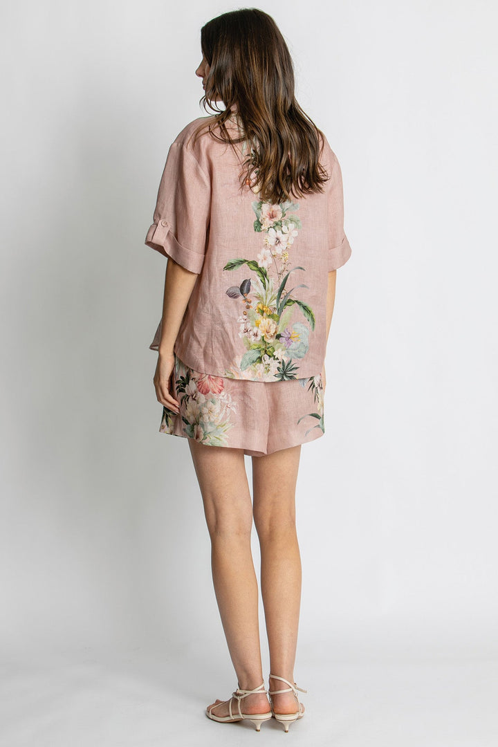 A woman shown from the back wearing a dusty pink linen short-sleeve top and matching shorts set with a colorful floral print, paired with beige strappy sandals against a light gray studio background.