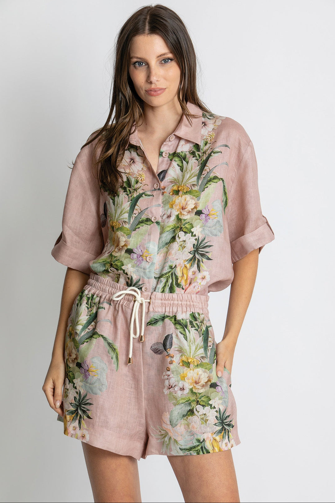 A woman modeling a dusty pink short-sleeve romper with a tropical floral print, featuring green leaves, yellow and white flowers, and a white drawstring waist.