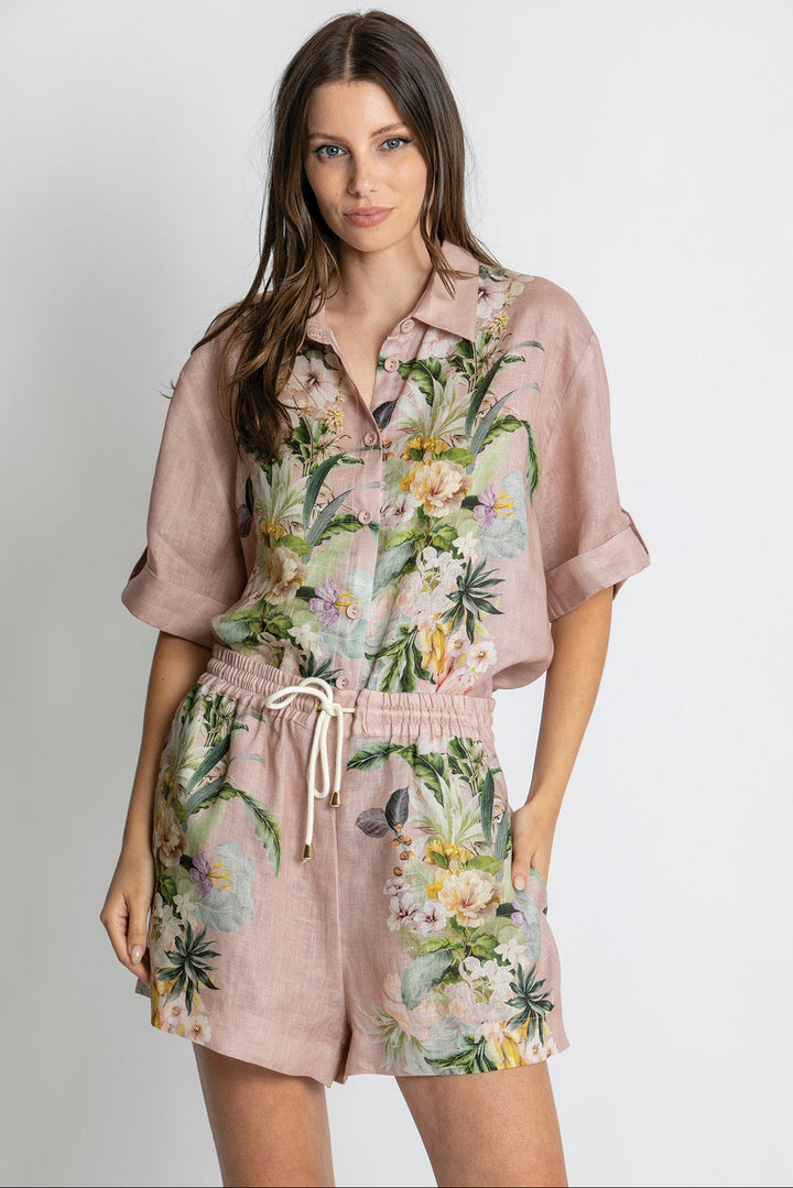 A woman modeling a dusty pink short-sleeve romper with a tropical floral print, featuring green leaves, yellow and white flowers, and a white drawstring waist.
