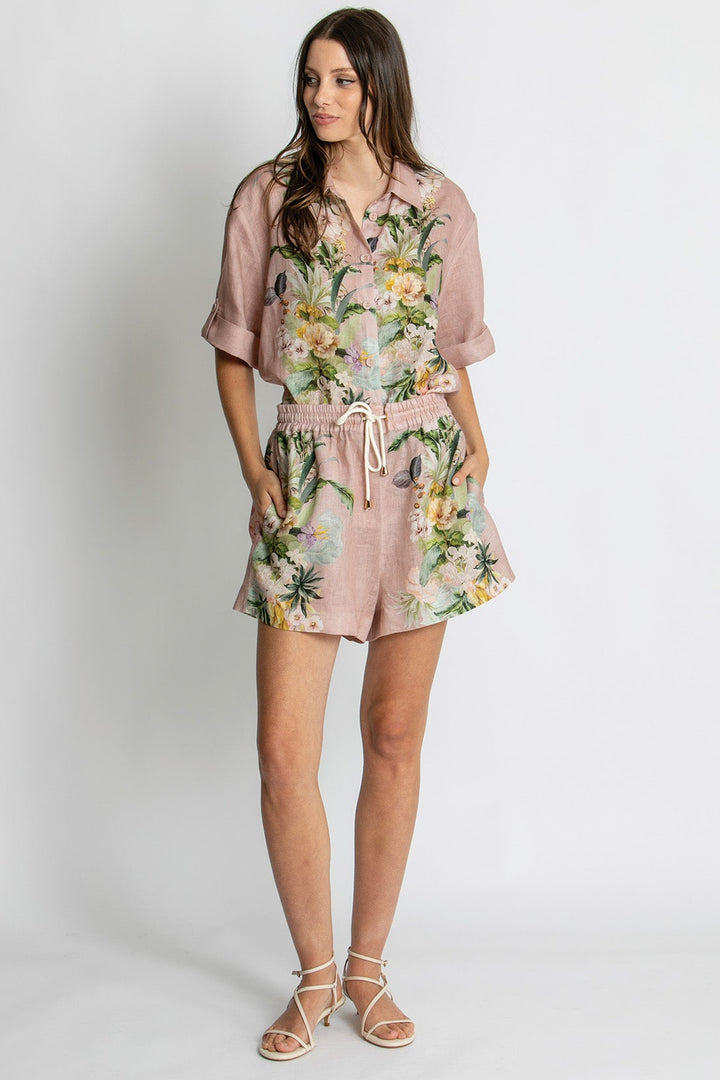 A woman modeling a dusty pink short-sleeve romper with a tropical floral print, featuring a button-up front, tie waist, and flared shorts, set against a plain light gray studio background.