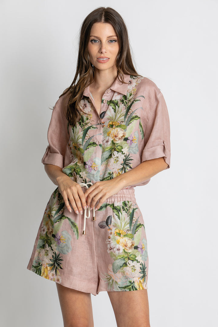 A woman modeling a dusty pink short-sleeve shirt and shorts set with a tropical floral print, featuring a button-down shirt top and matching drawstring shorts, against a light gray studio background.