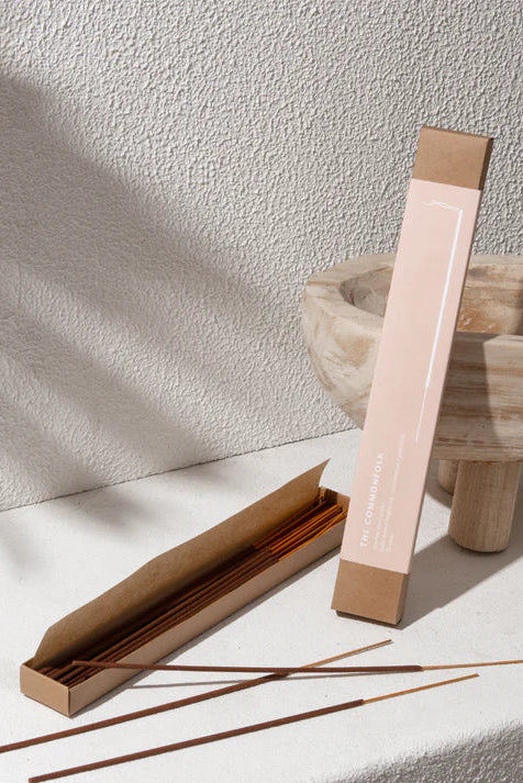 Incense sticks in a cardboard holder on a light surface with a wooden bowl in the background.