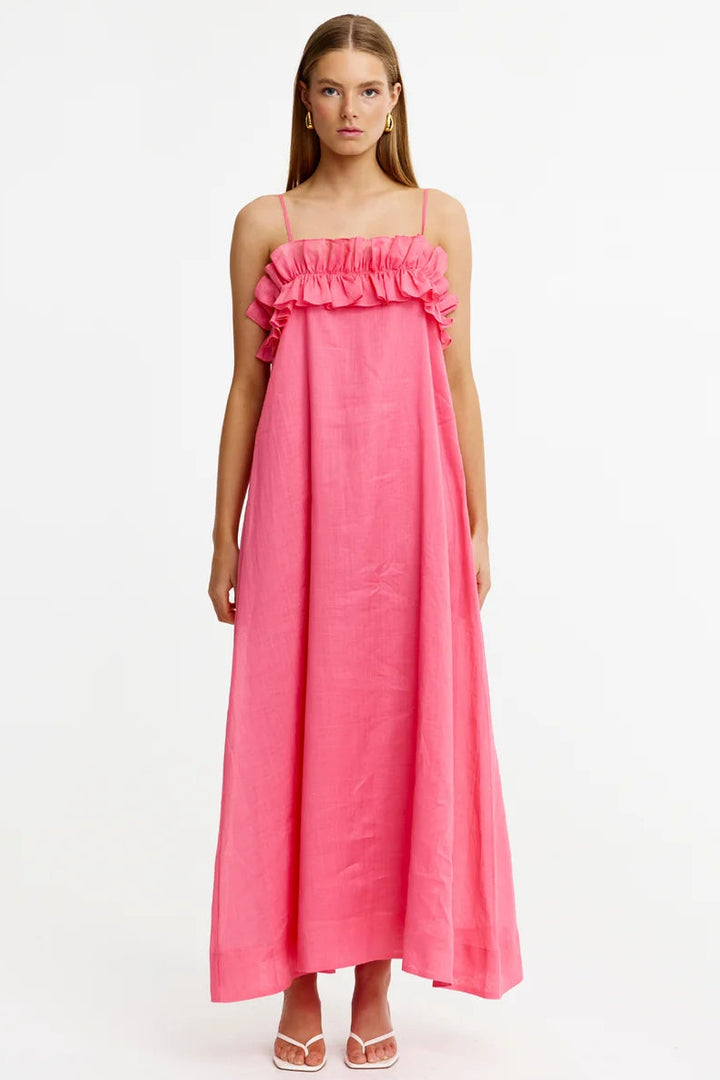 A woman modeling a bright pink off-the-shoulder maxi dress with a ruffled neckline, photographed against a plain white studio background.