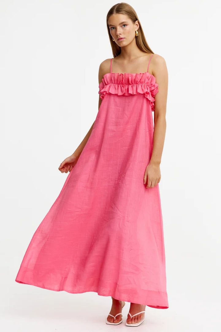 A woman modeling a bright pink maxi dress with thin straps, a ruffled neckline, and a flowing skirt, photographed against a plain white studio background.