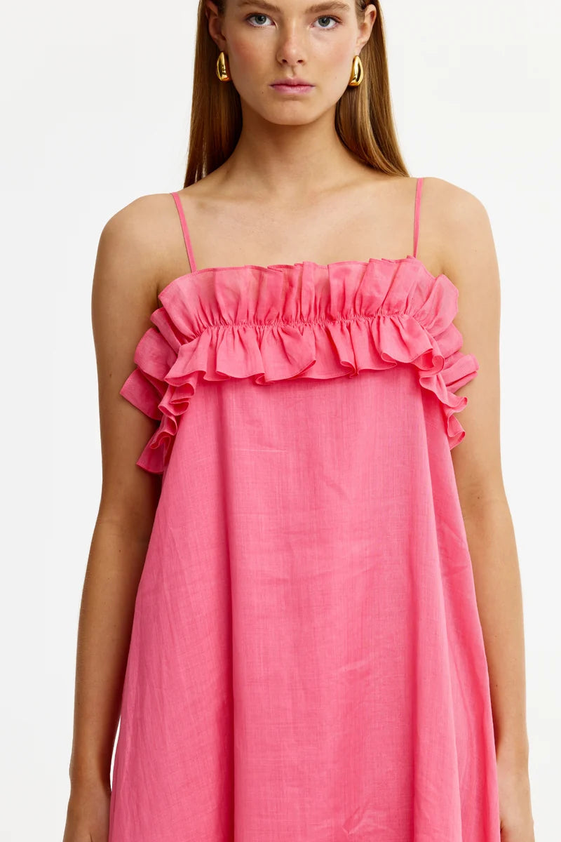 A woman wearing a bright pink sleeveless dress with thin straps and a ruffled neckline, shown against a plain light background.