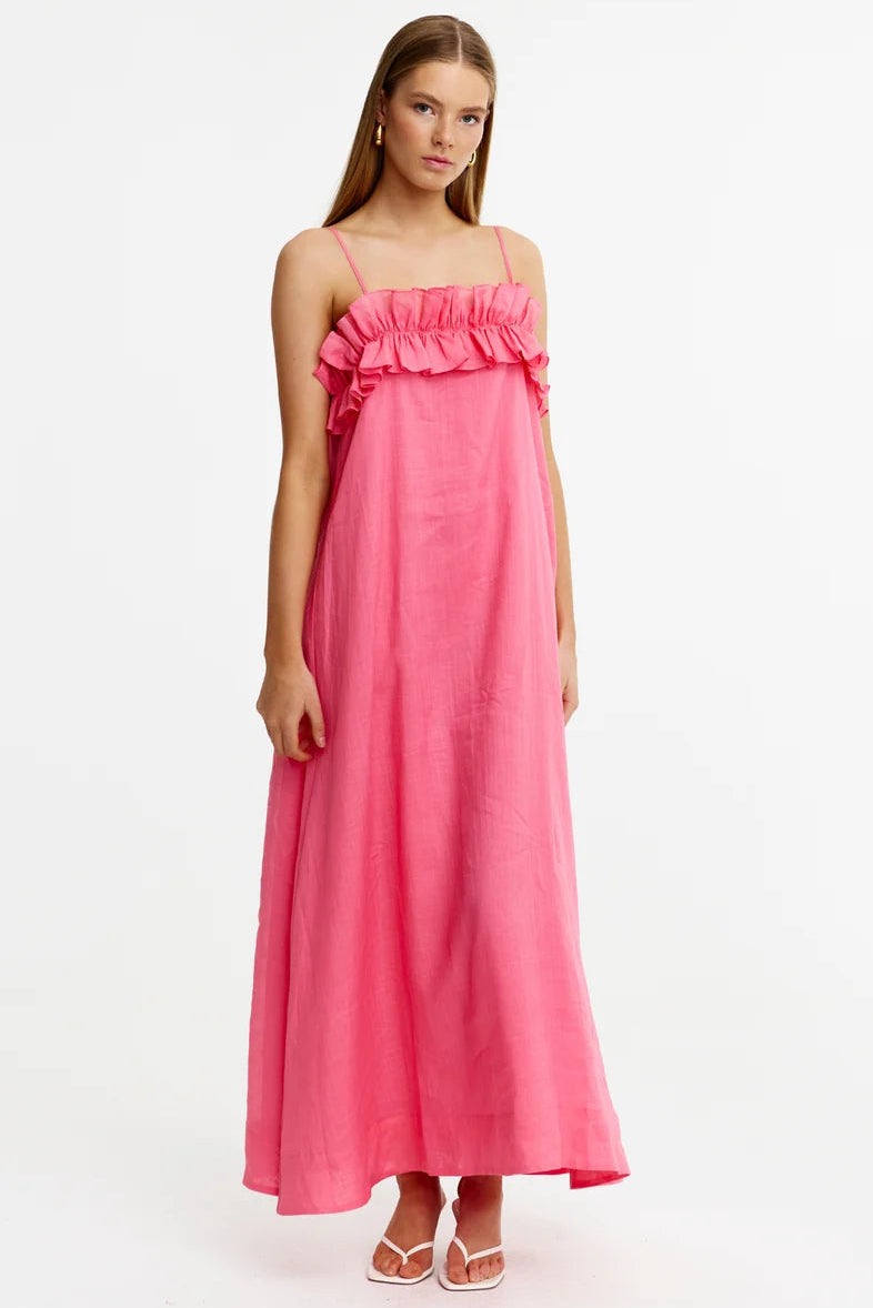 A woman modeling a bright pink maxi dress with thin straps, a ruffled off-the-shoulder neckline, and a flowing skirt, standing against a plain white studio background.