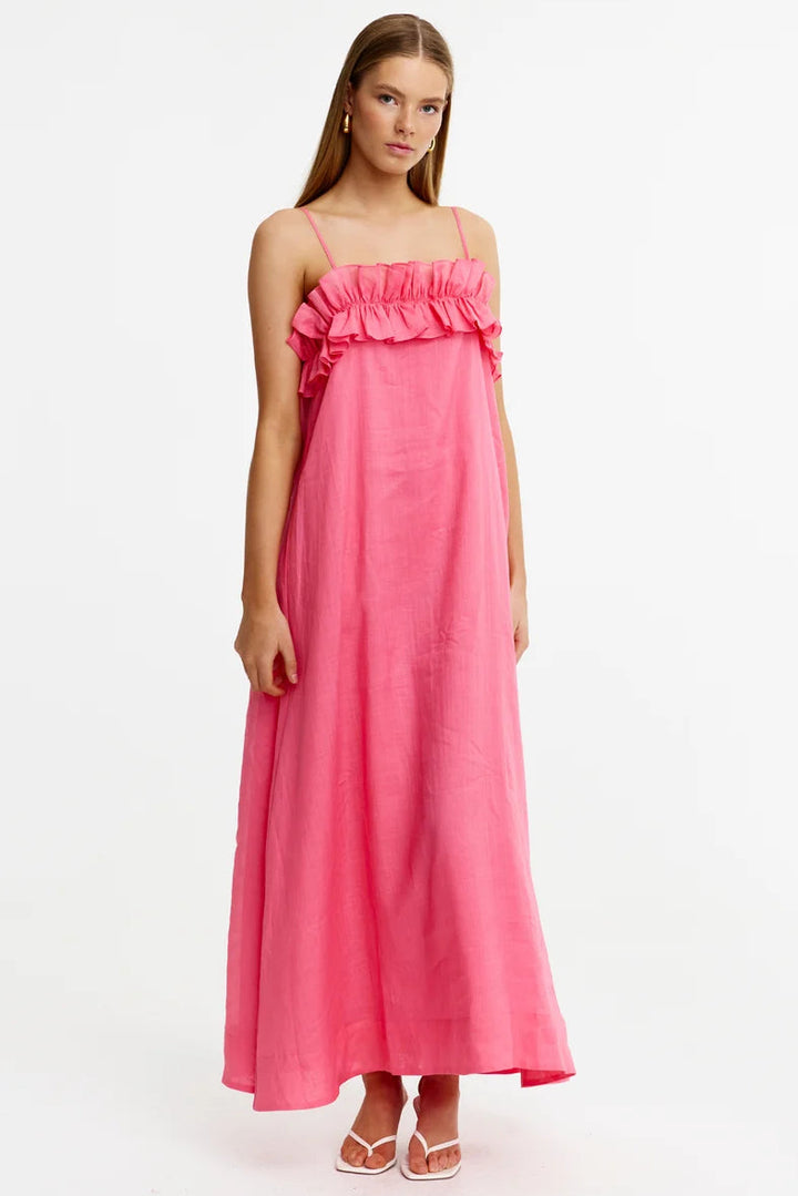 A woman modeling a bright pink maxi dress with thin straps, a ruffled off-the-shoulder neckline, and a flowing skirt, standing against a plain white studio background.