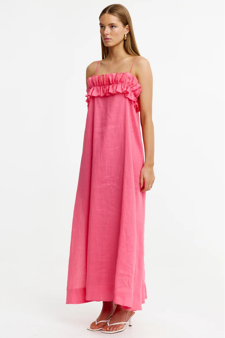 A woman modeling a bright pink, strapless maxi dress with a ruffled neckline, standing against a plain white studio background.