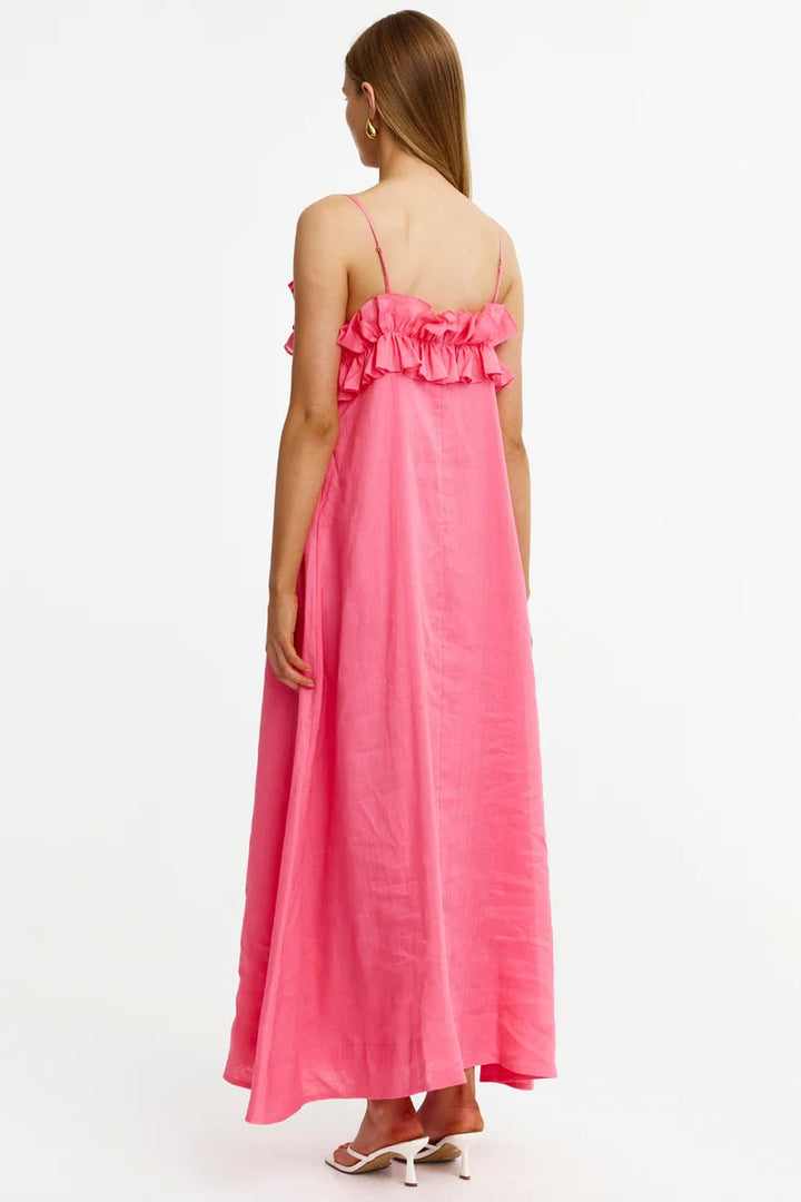 A woman viewed from the back wearing a bright pink maxi dress with thin straps, a ruffled off-the-shoulder neckline, and a flowing skirt, paired with white heeled sandals against a plain white studio background.