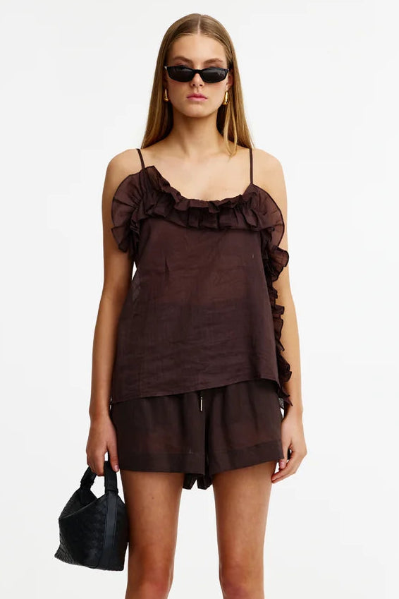 A model stands against a plain white studio background wearing a dark brown ruffled camisole top and matching shorts, with black sunglasses, sandals, and a small black handbag.