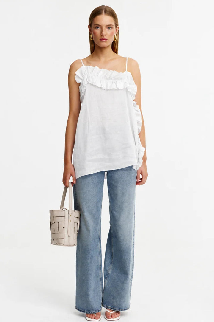 A model stands against a plain white studio background wearing a white ruffled spaghetti-strap top, wide-leg light-wash blue jeans, and holding a small beige woven tote bag.