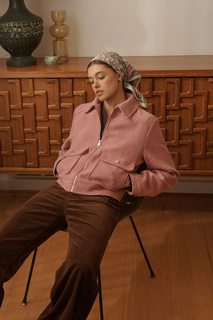 Woman wearing a pink jacket and brown pants sitting on a chair in a room with wooden furniture.