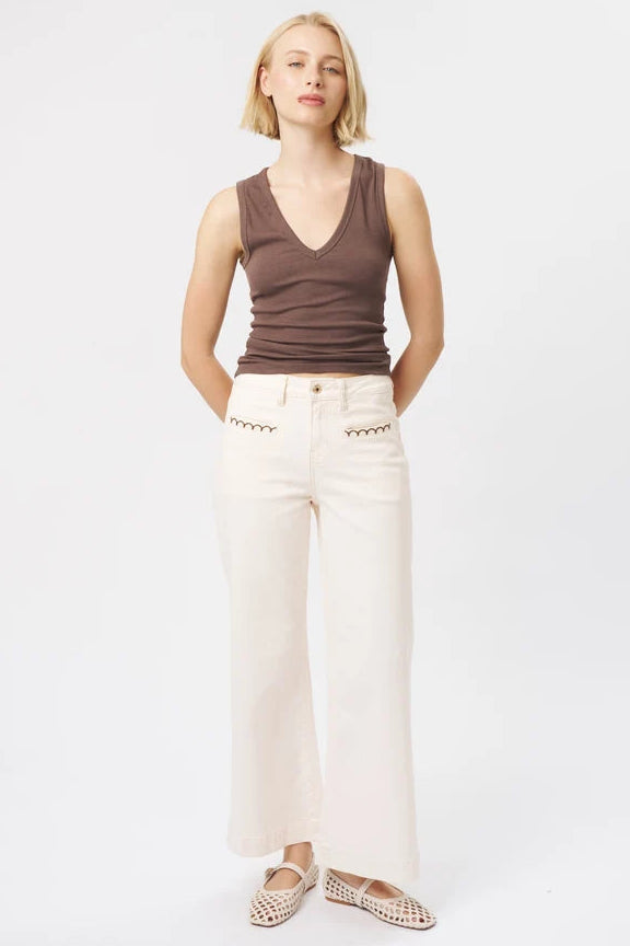 A woman stands against a plain white studio background wearing a brown ribbed sleeveless V-neck top, off-white wide-leg pants, and beige woven flats.