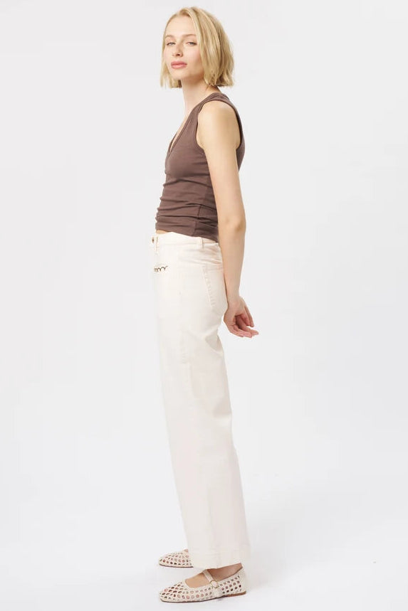 A woman stands in profile against a plain white studio background, wearing a fitted brown sleeveless top, off-white wide-leg pants, and beige woven flats.