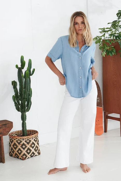 A woman stands barefoot in a bright indoor room wearing a light blue short-sleeve button-front shirt and white wide-leg pants. A potted cactus, wooden bench, cabinet, and plant are visible in the background.