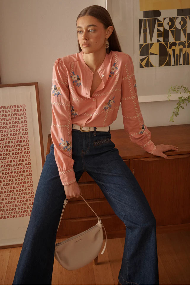 Woman in a pink blouse and blue jeans holding a beige handbag in a room with a wooden desk and framed pictures.