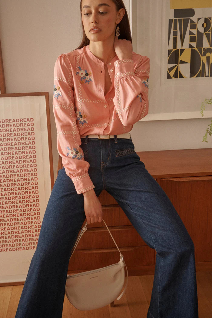 Woman wearing a pink embroidered blouse and blue jeans, sitting indoors.