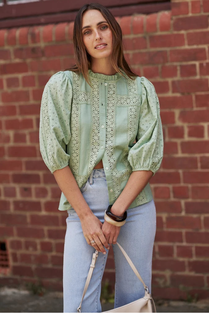A woman wearing a sage green eyelet blouse with puff sleeves and light blue jeans, standing in front of a red brick wall and holding a cream-colored handbag.