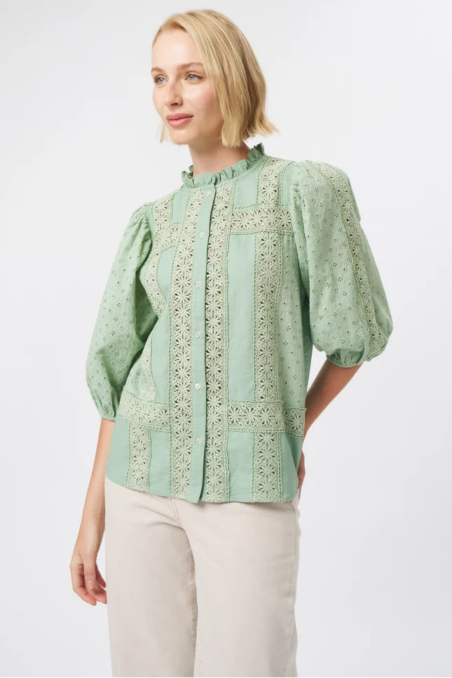 A woman modeling a sage green blouse with intricate floral embroidery and puff sleeves, paired with light beige trousers, against a plain light gray background.