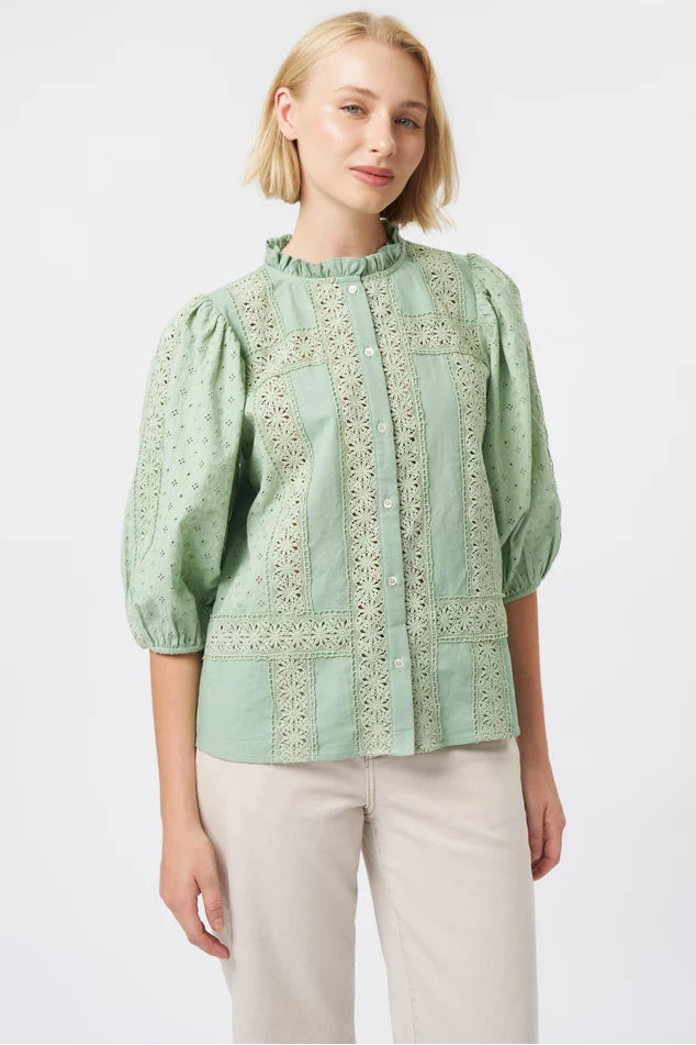 A woman modeling a light sage green blouse with intricate embroidery and puff sleeves, paired with beige trousers, against a plain white studio background.