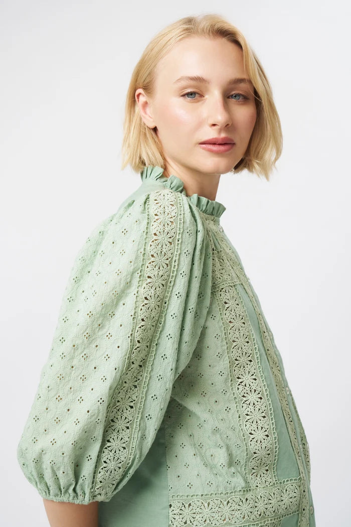 A blonde woman wearing a sage green eyelet blouse with puff sleeves and a ruffled high neck, photographed against a clean light gray studio background.