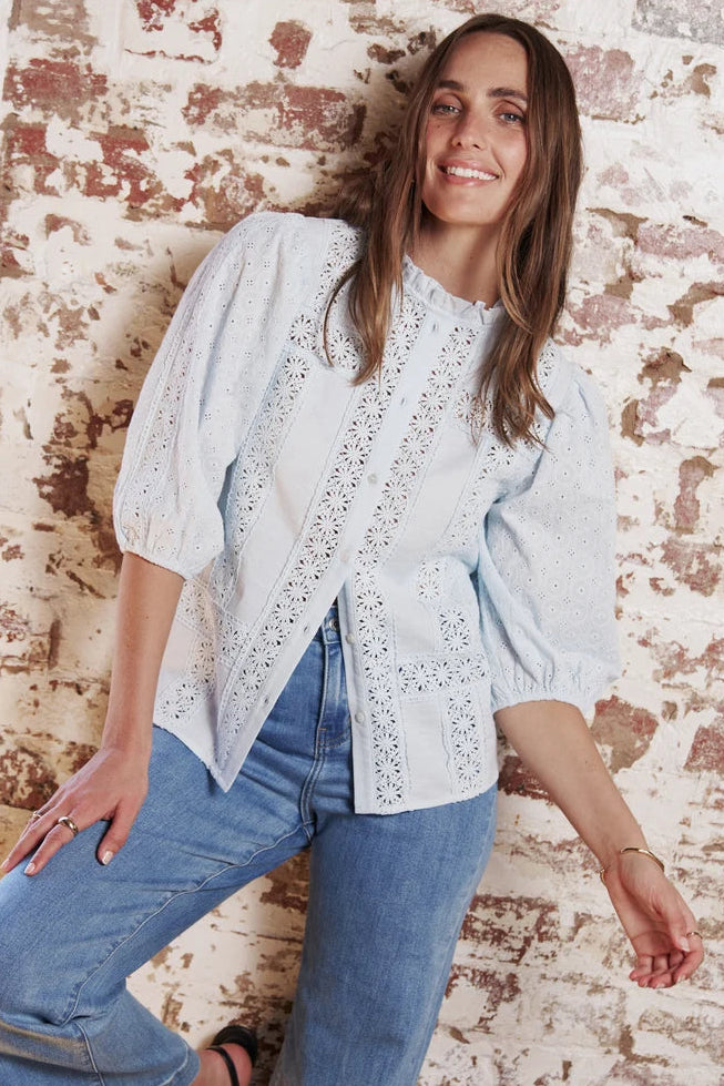 A woman wearing a light blue eyelet blouse with puff sleeves and blue jeans, leaning against a distressed brick wall.