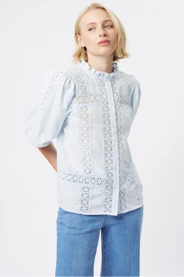 A woman modeling a light blue eyelet blouse with puff sleeves and a ruffled high collar, paired with blue jeans, against a plain white studio background.