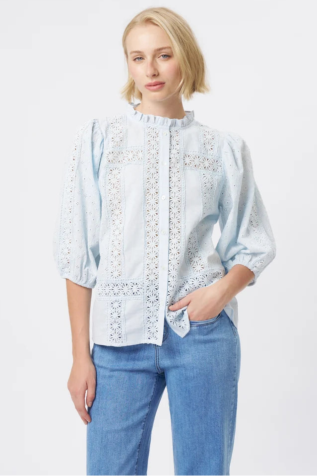 A woman modeling a light blue eyelet blouse with three-quarter sleeves and a ruffled neckline, paired with blue jeans, against a plain light gray studio background.