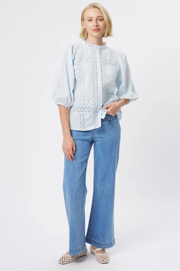 A woman modeling a light blue eyelet blouse with puff sleeves and a high ruffled collar, paired with wide-leg blue jeans and beige woven sandals, against a plain white studio background.